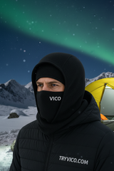 Person wearing a black VICO branded jacket and balaclava in a snowy landscape with a tent and northern lights.